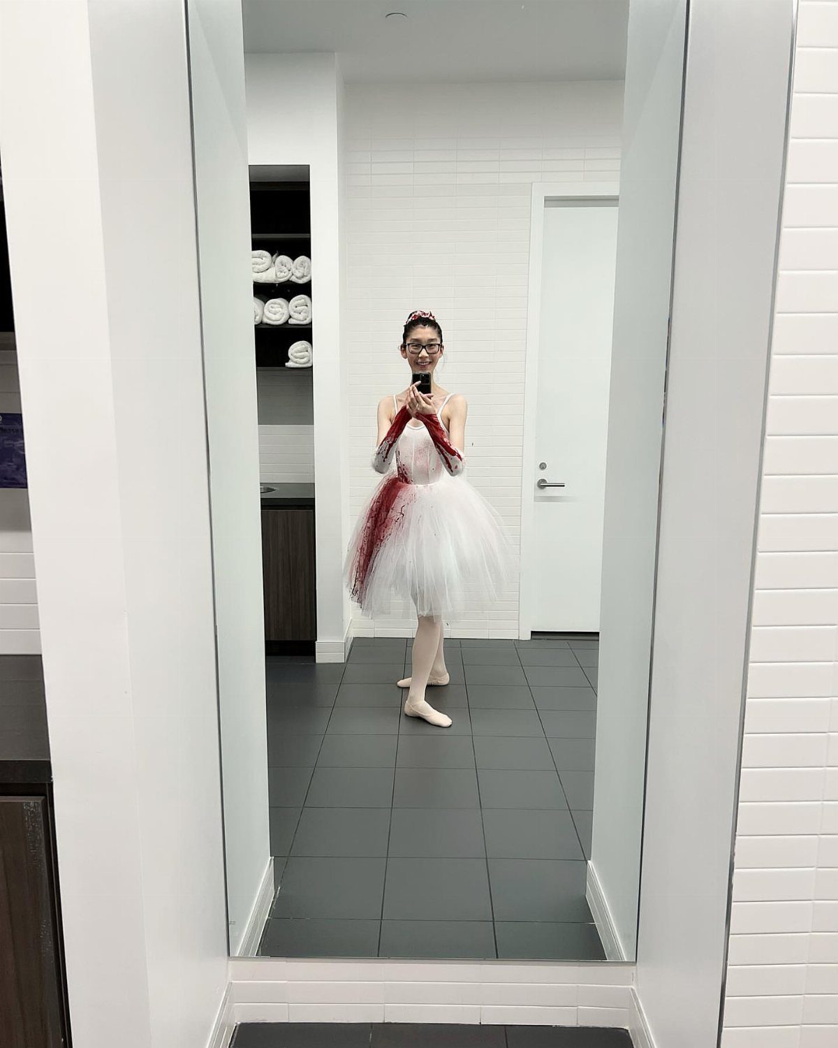 Person in a blood-stained white ballerina tutu and ballet shoes taking a mirror selfie.