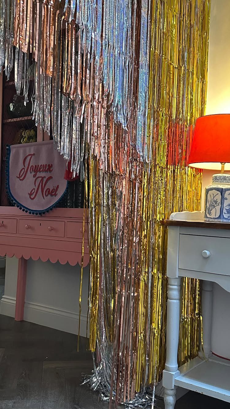 22 Christmas Party Themes for Adults Ideas (2026) 18 Gold and silver tinsel curtain backdrop next to a pink cabinet and red lamp.