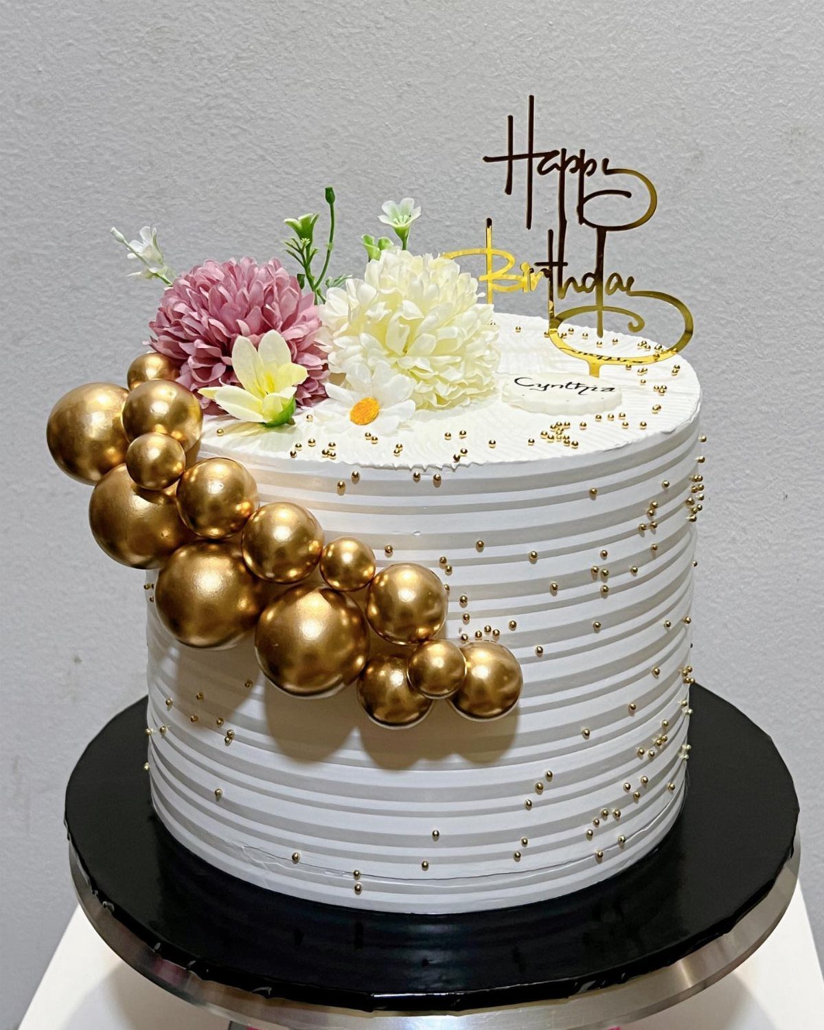 White ridged birthday cake decorated with gold spheres, sprinkles, and fresh flowers.