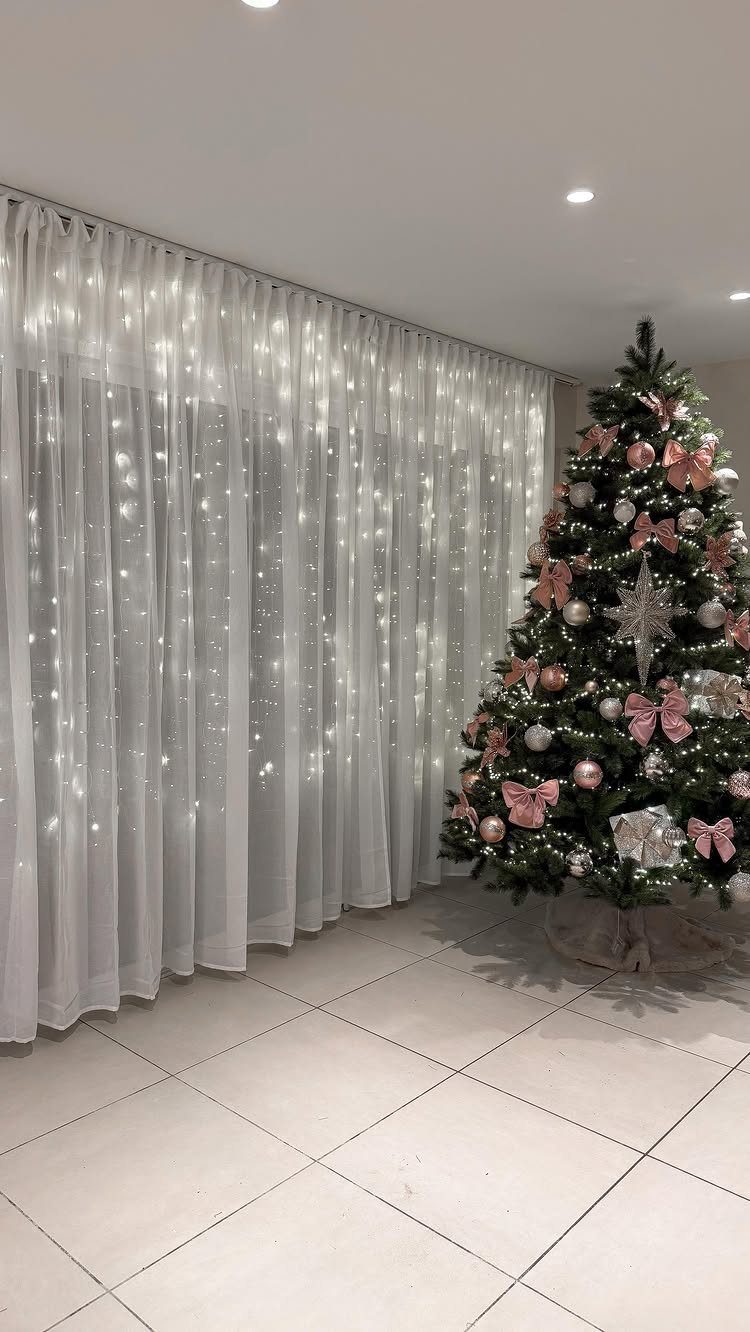 36 Christmas Decor Ideas for the Home: Doors, Tables, Trees & More 32 Decorated Christmas tree with pink bows beside white curtains with fairy lights.