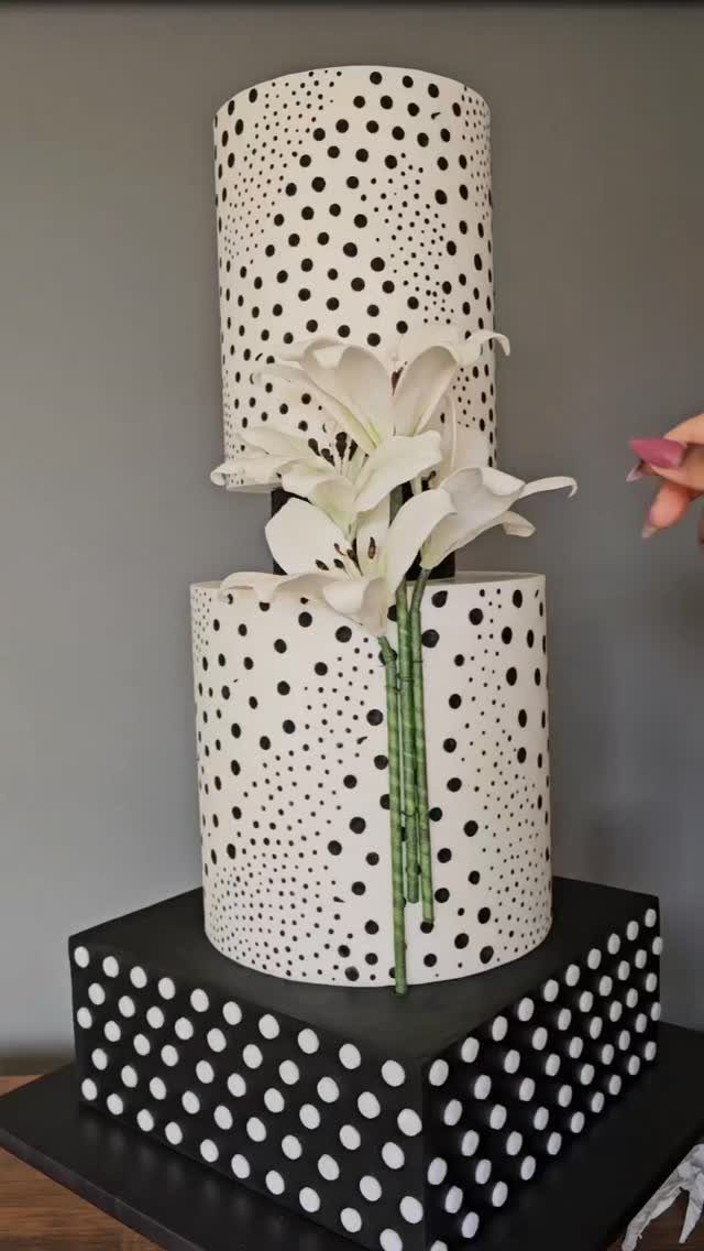 Two-tiered white cake with black polka dots and white lilies on a square base.